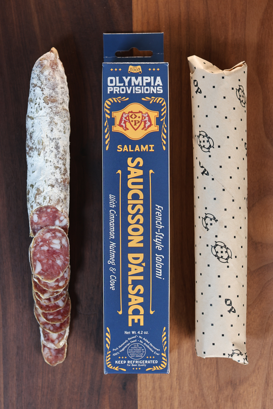 Olympia Provisions Saucisson d'Alsace with clove, nutmeg, allspice, & cinnamon, packaged in a blue box on a wood background. A partially sliced salami sits next to the box.