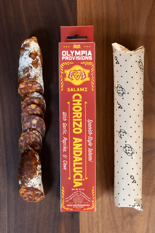 Olympia Provisions Chorizo Andalucia paprika, garlic, and clove, packaged in a red box on a wood background. A partially sliced salami sits next to the box.