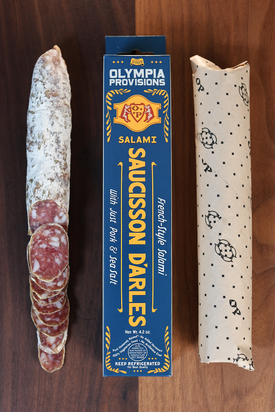 Olympia Provisions Saucisson d'Arles with sea salt, packaged in a blue box on a wood background. A partially sliced salami sits next to the box.