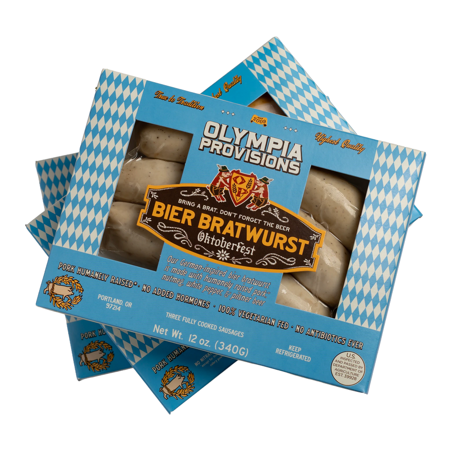 Three boxes of Olympia Provisions Bier Bratwurst sausages stacked on a transparent background. The packaging features a blue and white diamond pattern.
