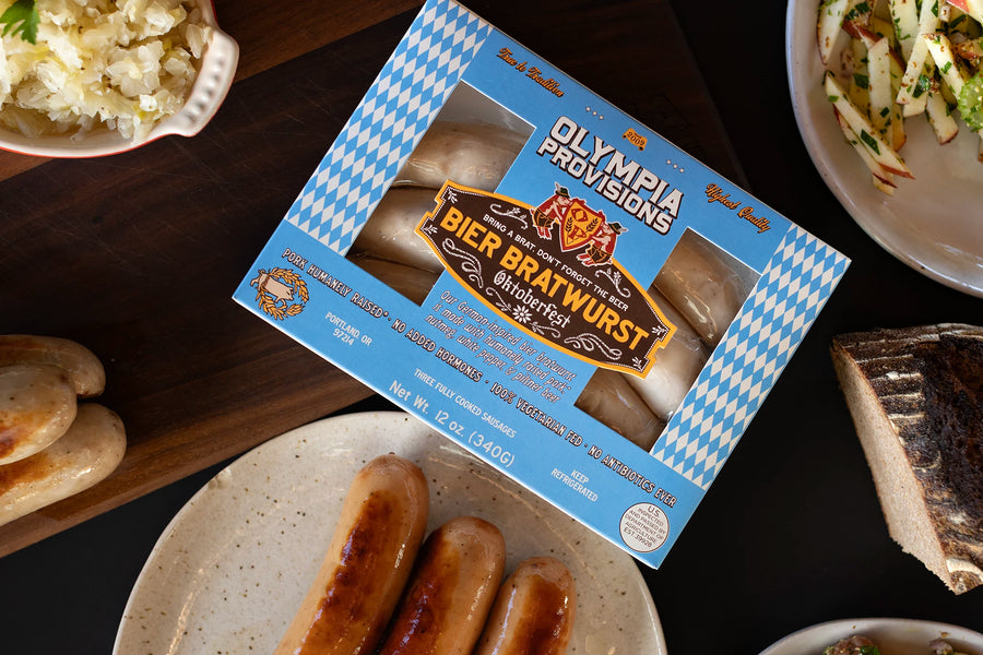 One pack of Olympia Provisions Bier Bratwurst sausages surrounded by plates of food on a wooden table.