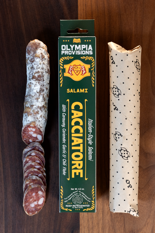 Olympia Provisions Cacciatore with caraway, coriander, dried red chili, & black pepper, packaged in a green box on a wood background. A partially sliced salami sits next to the box.