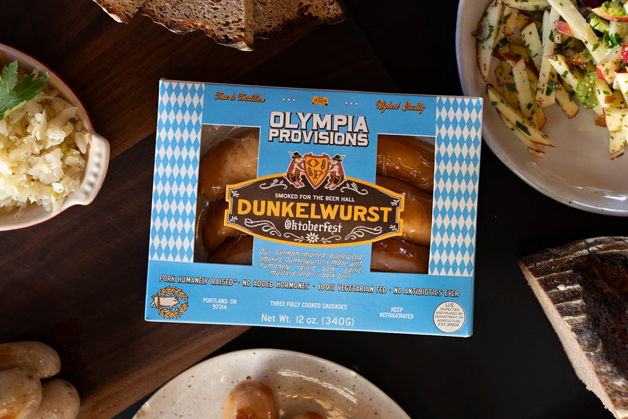 One pack of Olympia Provisions Dunkelwurst sausages surrounded by plates of food on a wooden table.