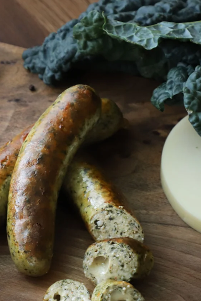 Three cooked sausage links are arranged on a wooden cutting board alongside kale and a round slice of provolone cheese.