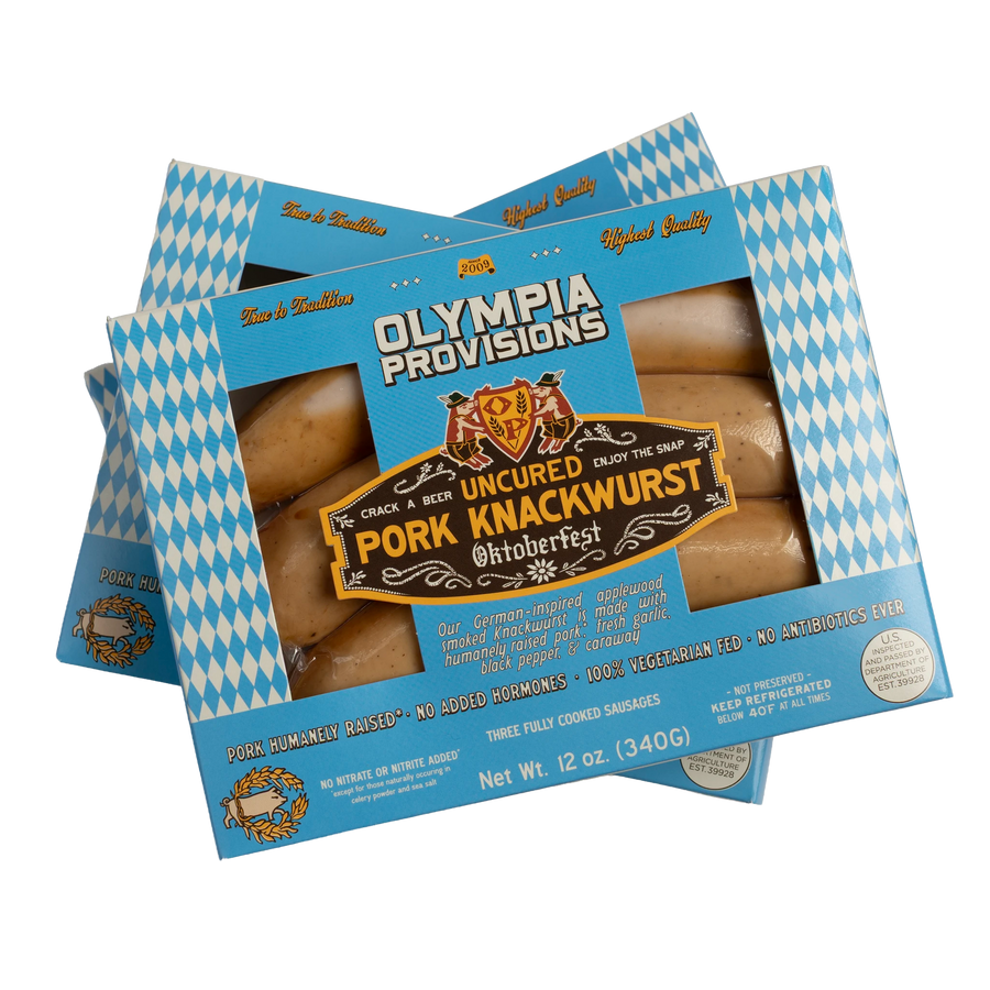 Three boxes of Olympia Provisions Pork Knackwurst sausages stacked on a transparent background. The packaging features a blue and white diamond pattern.