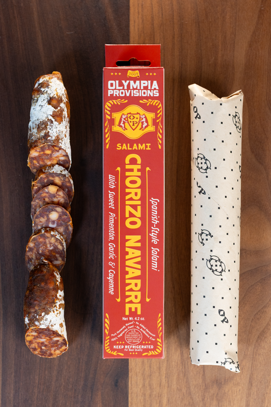 Olympia Provisions Chorizo Navarre with sweet paprika, garlic & cayenne, packaged in a red box on a wood background. A partially sliced salami sits next to the box.