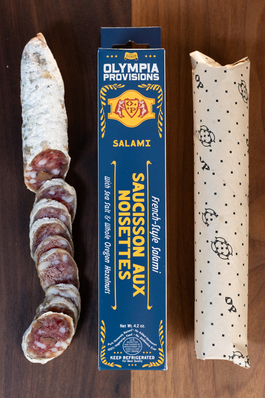 Olympia Provisions Saucisson aux Noisette with Oregon hazelnuts, packaged in a blue box on a wood background. A partially sliced salami sits next to the box.