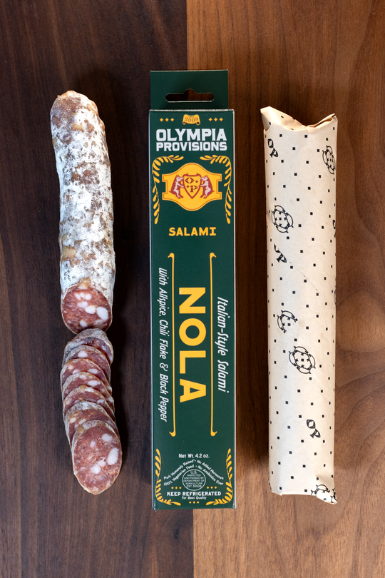 Olympia Provisions Nola with sweet and black pepper, chili flake & allspice, packaged in a green box on a wood background. A partially sliced salami sits next to the box.