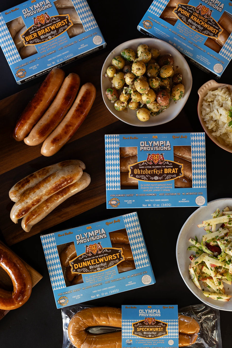 Olympia Provisions Oktoberfest sausages sausages surrounded by plates of food on a wooden table. One pack each of Bier Bratwurst, Oktoberfest Brat, Dunkelwurst, Pork Knackwurst, and Speckwurst are visible.