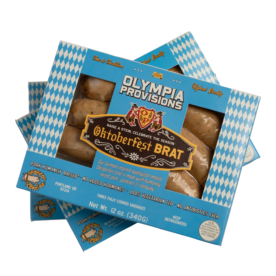 Three boxes of Olympia Provisions Oktoberfest Brat sausages stacked on a transparent background. The packaging features a blue and white diamond pattern.