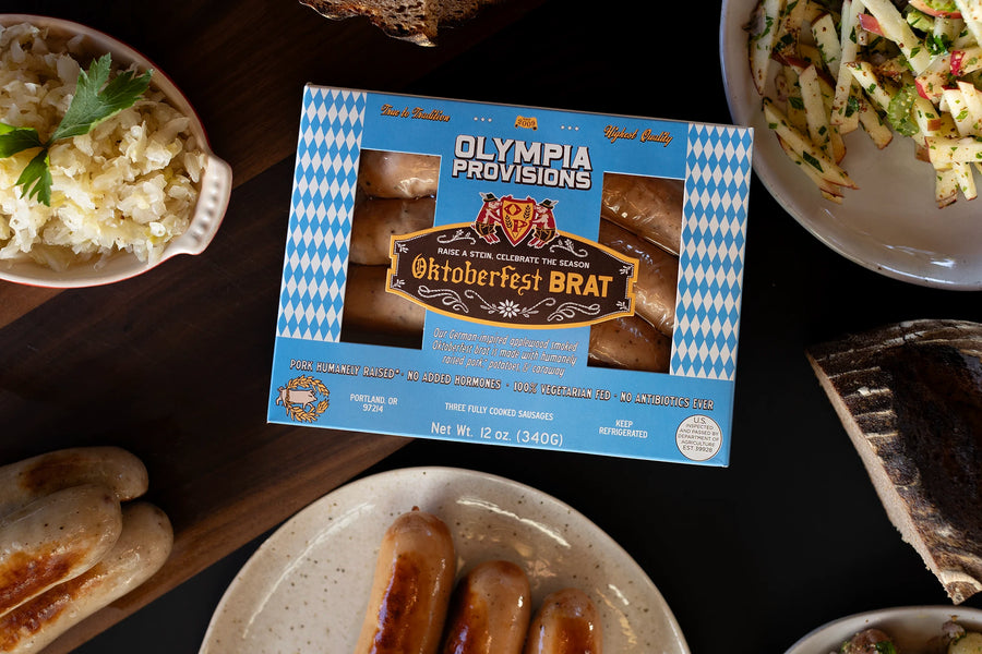 One pack of Olympia Provisions Oktoberfest Brat sausages surrounded by plates of food on a wooden table.