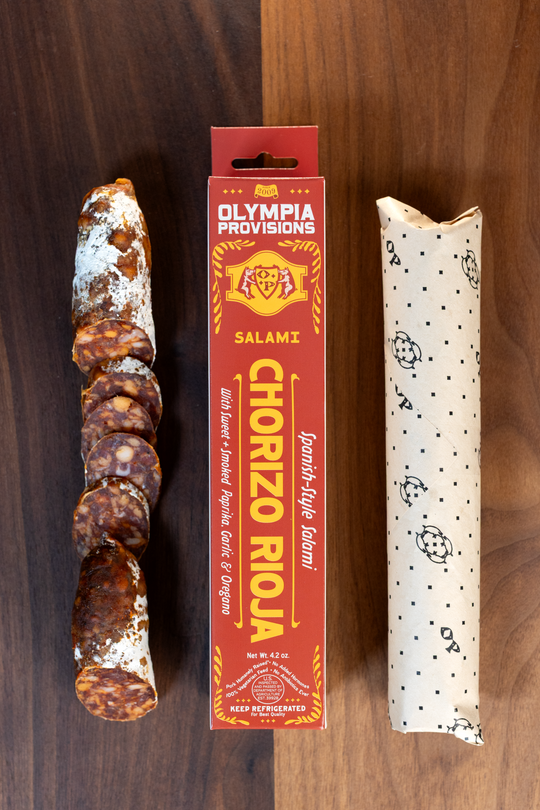 Olympia Provisions Chorizo Rioja with sweet and smoked paprika, fresh garlic, and oregano, packaged in a red box on a wood background. A partially sliced salami sits next to the box.