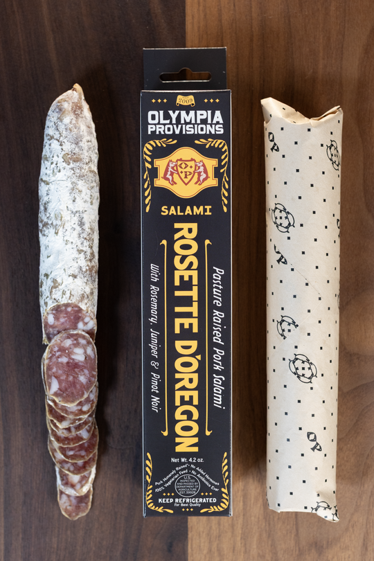 Olympia Provisions Rosette d'Oregon with rosemary, juniper, and pinot noir, packaged in a black box on a wood background. A partially sliced salami sits next to the box.