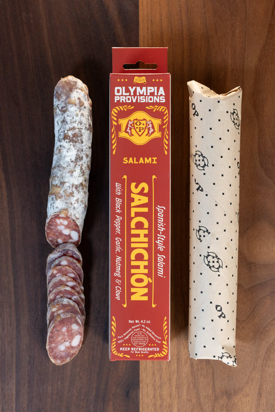 Olympia Provisions Salchichon with cinnamon, clove, nutmeg, black pepper, garlic, & paprika packaged in a red box on a wood background. A partially sliced salami sits next to the box.