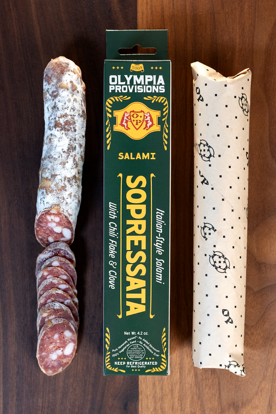 Olympia Provisions Sopressata with clove, oregano, garlic, & chili flake, packaged in a green box on a wood background. A partially sliced salami sits next to the box.