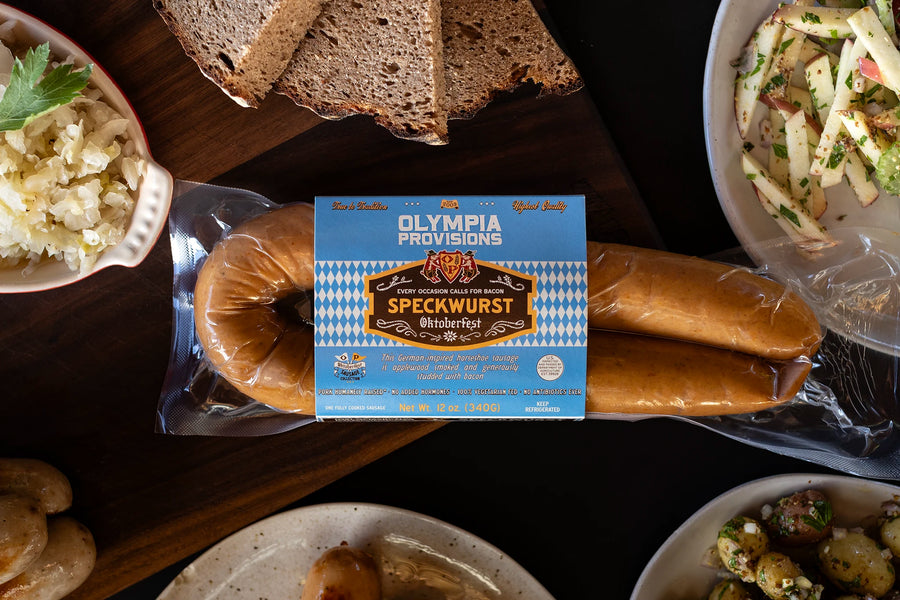One pack of Olympia Provisions Speckwurst sausages surrounded by plates of food on a wooden table.