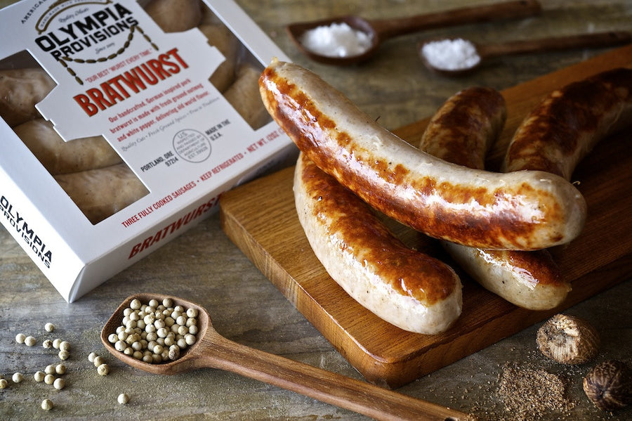 Grilled bratwursts on a wooden cutting board next to a package of Olympia Provisions bratwurst. The scene includes wooden spoons filled with white peppercorns and other spices on a rustic wooden surface