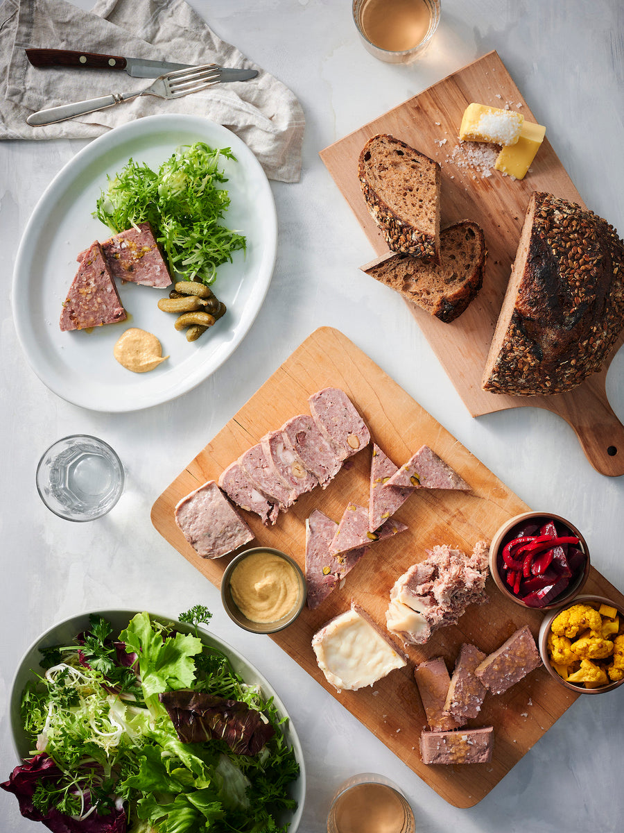 Pates on a cutting board with pickles, bread, and a bowl of salad nearby.